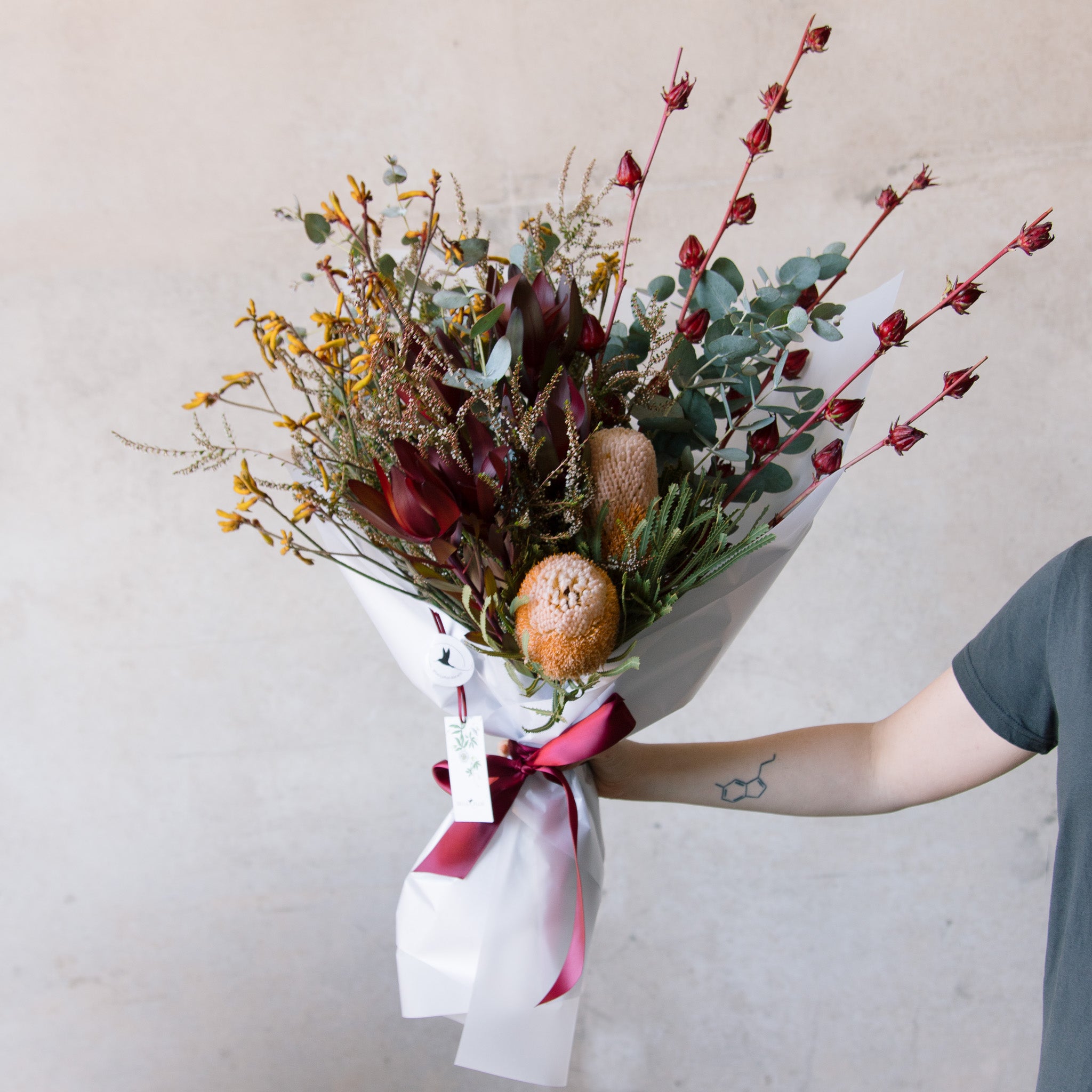 A bouquet of Australian native flowers with a mix of earthy colours, predominantly red and yellow, wrapped in white paper with a red ribbon.