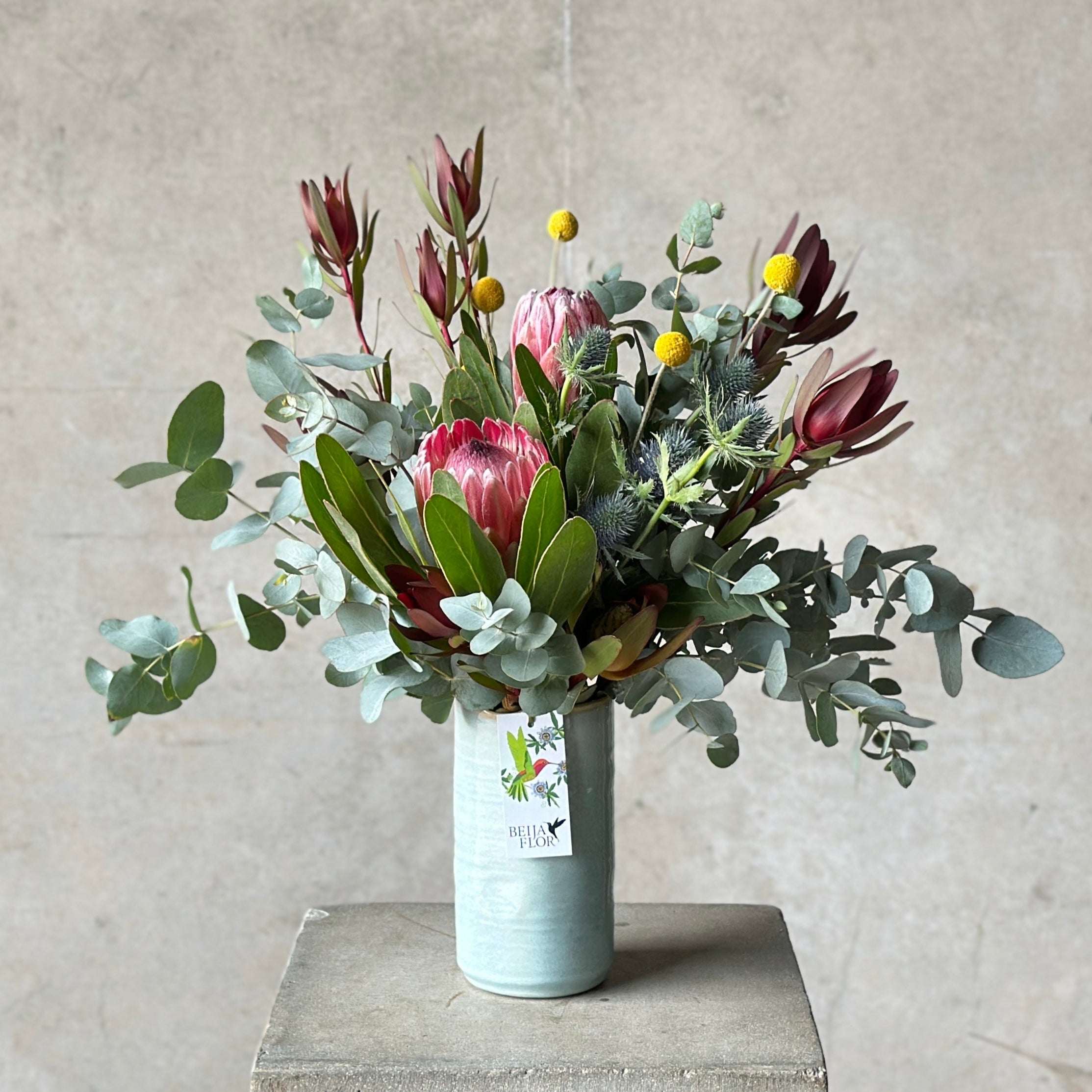 A floral arrangement featuring a variety of native Australian flowers such as proteas and banksias, with green foliage and decorative elements in a blue glazed vase.